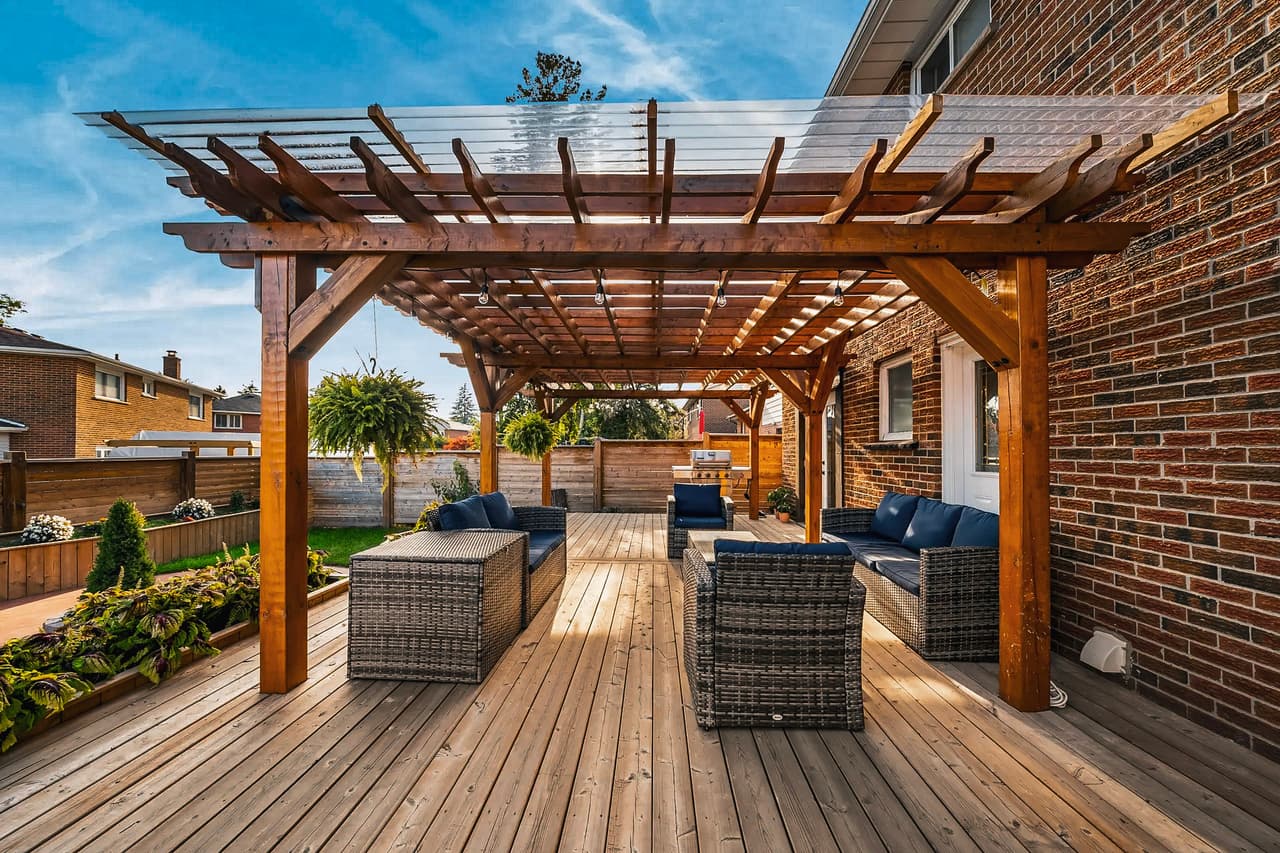Grande terrasse en bois attenante à une maison en briques, abritée par une pergola moderne et meublée de canapés en rotin