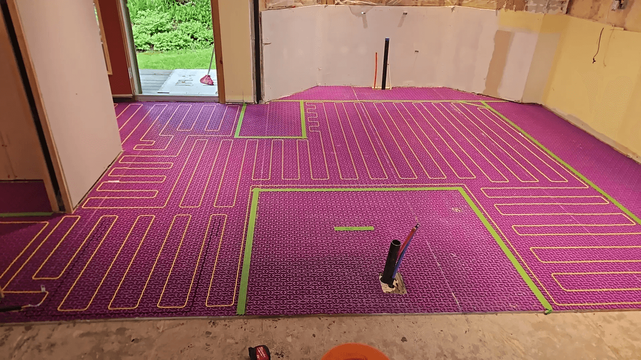 Installation de plancher chauffant hydronique avec membrane de désolidarisation mauve, tuyaux rouges, ruban vert et sorties de plomberie dans une pièce en rénovation