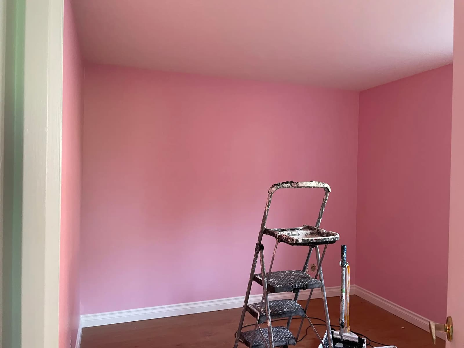 Chambre en cours de rénovation avec murs fraîchement peints en rose, plinthes blanches, sol en bois et matériel de peinture.