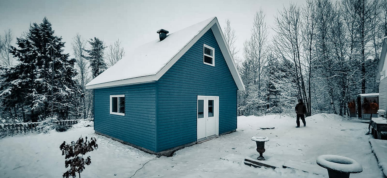 Petite maison en bois bleue avec un toit couvert de neige, située dans une forêt hivernale. Cadre paisible avec des arbres enneigés et une personne en arrière-plan.