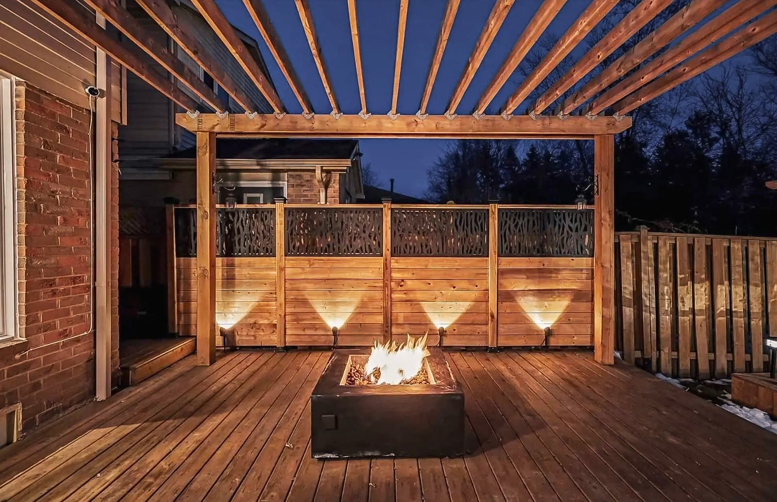 Terrasse en bois avec pergola et foyer extérieur illuminé, entourée d’une clôture décorative en bois, idéale pour les soirées chaleureuses à l’extérieur.