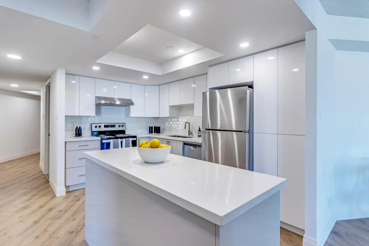 Cuisine de style contemporain à Montréal, mettant en valeur un îlot central blanc avec un comptoir en quartz. Les armoires blanches brillantes et les appareils électroménagers en acier inoxydable apportent une touche d'élégance à l'espace. Le sol en bois clair et l'éclairage encastré créent une ambiance lumineuse et accueillante