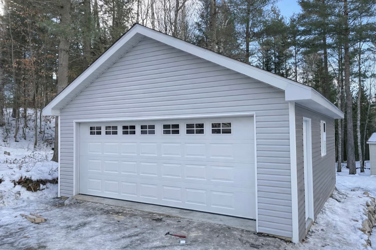 Garage détaché moderne avec revêtement de vinyle gris, grande porte de garage blanche, soffites et fascias blancs, toiture à deux versants en environnement boisé enneigé.