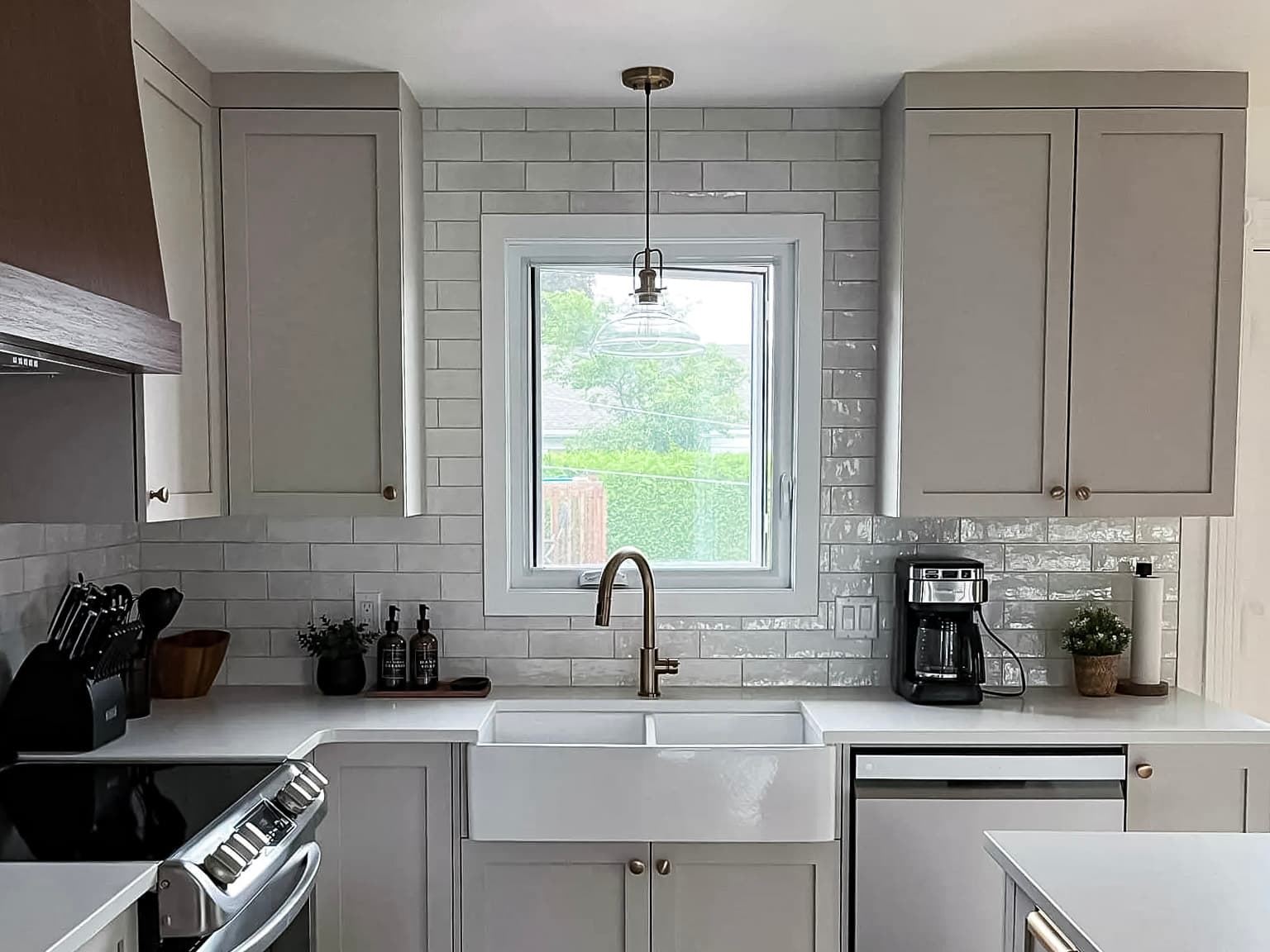 Cuisine moderne avec armoires grises, comptoirs blancs, évier de style farmhouse et électroménagers en acier inoxydable, éclairée par une suspension en verre.