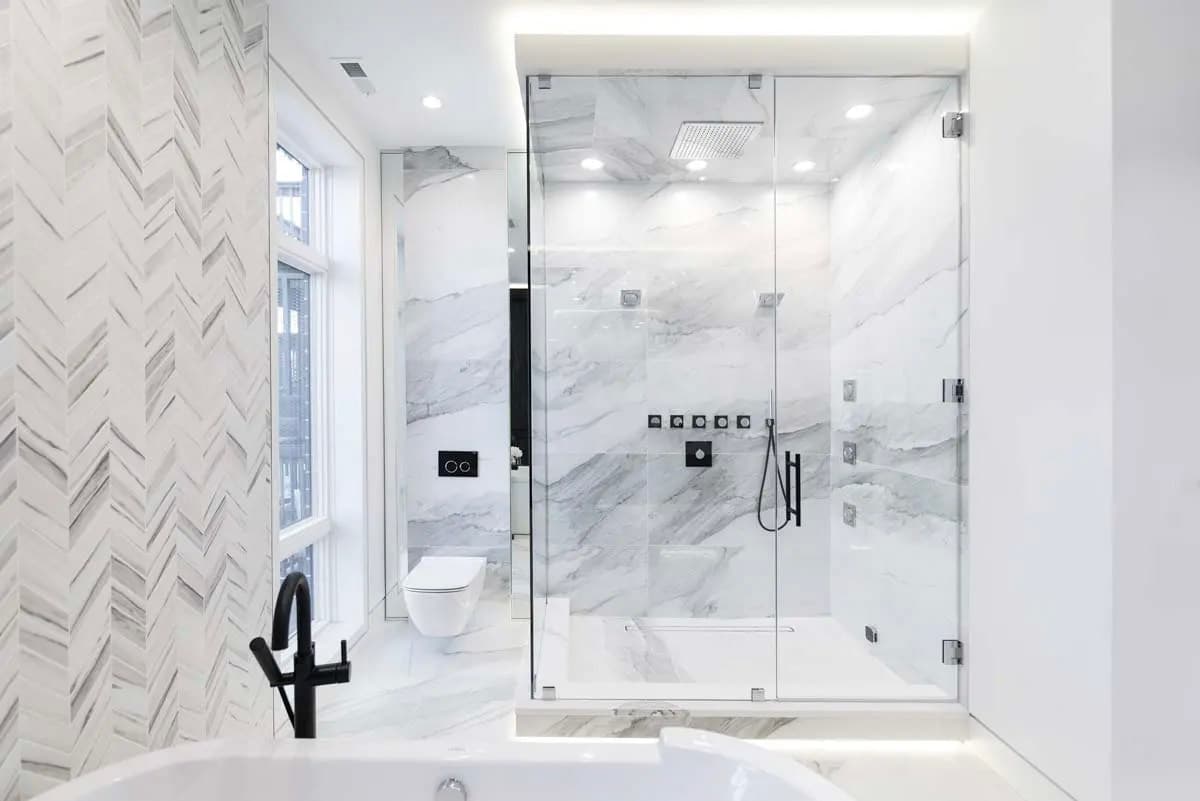Salle de bain moderne et luxueuse avec douche vitrée, murs en marbre blanc, carrelage chevron, robinetterie noire, baignoire blanche et éclairage encastré pour un design épuré et lumineux
