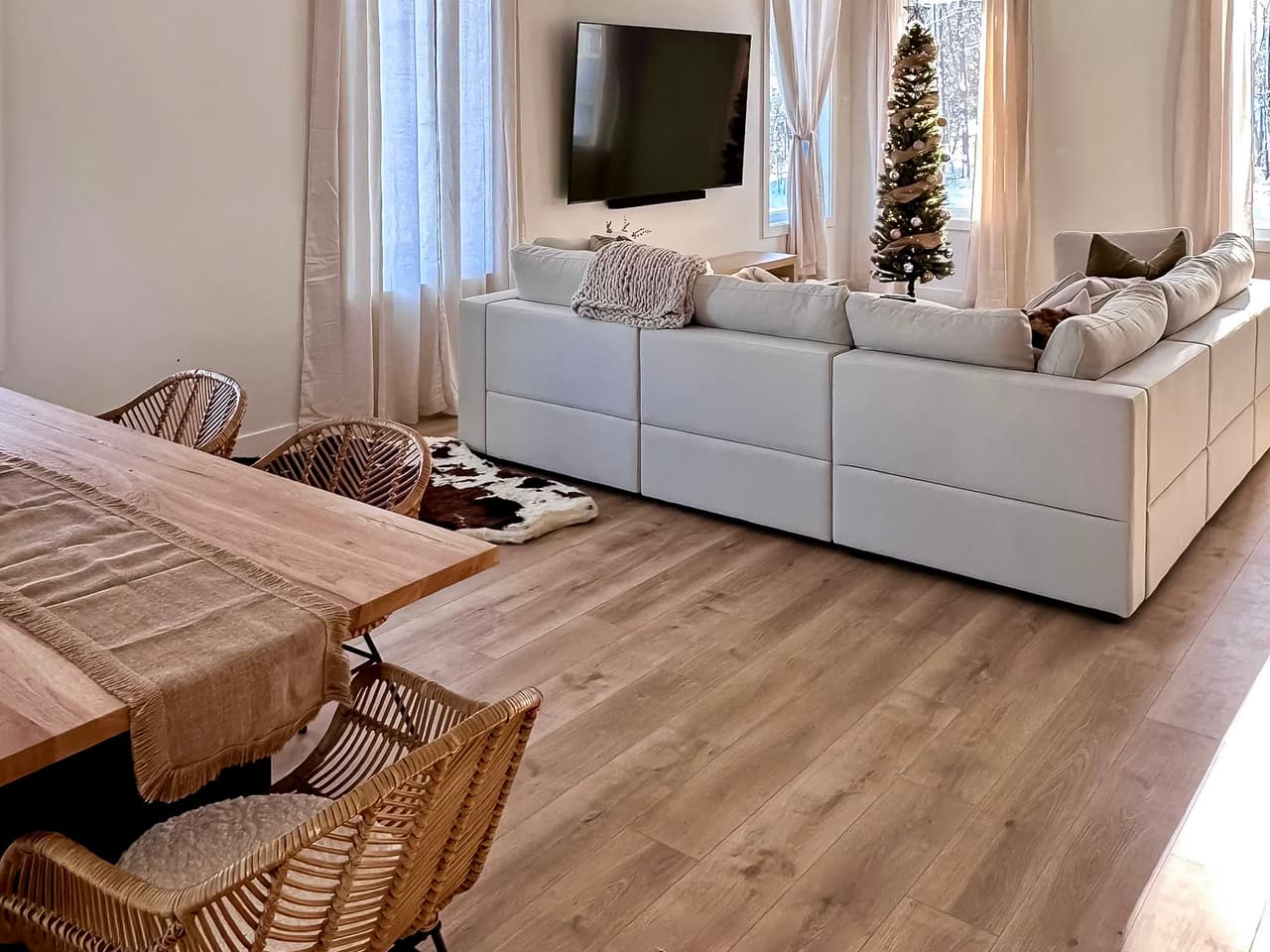 Salon lumineux avec canapé sectionnel blanc, rideaux beiges transparents, téléviseur mural, table à manger en bois