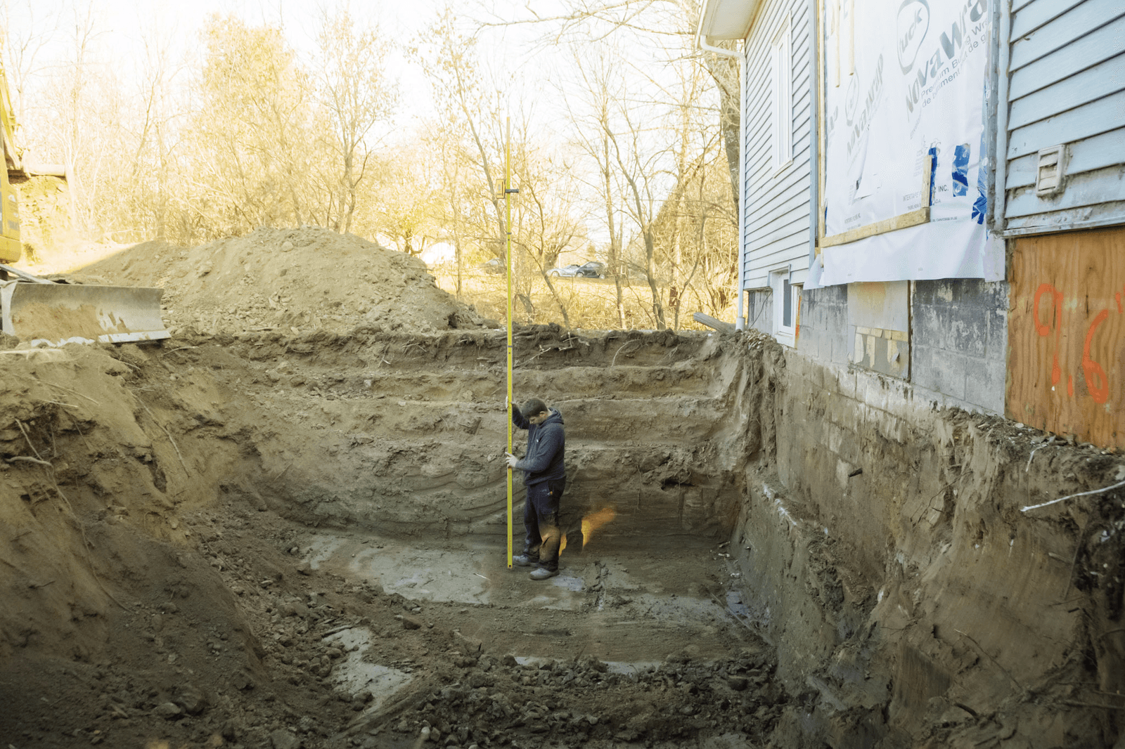 Ouvrier mesurant la profondeur d’une excavation de fondation à l’aide d’une perche graduée près d’une maison.