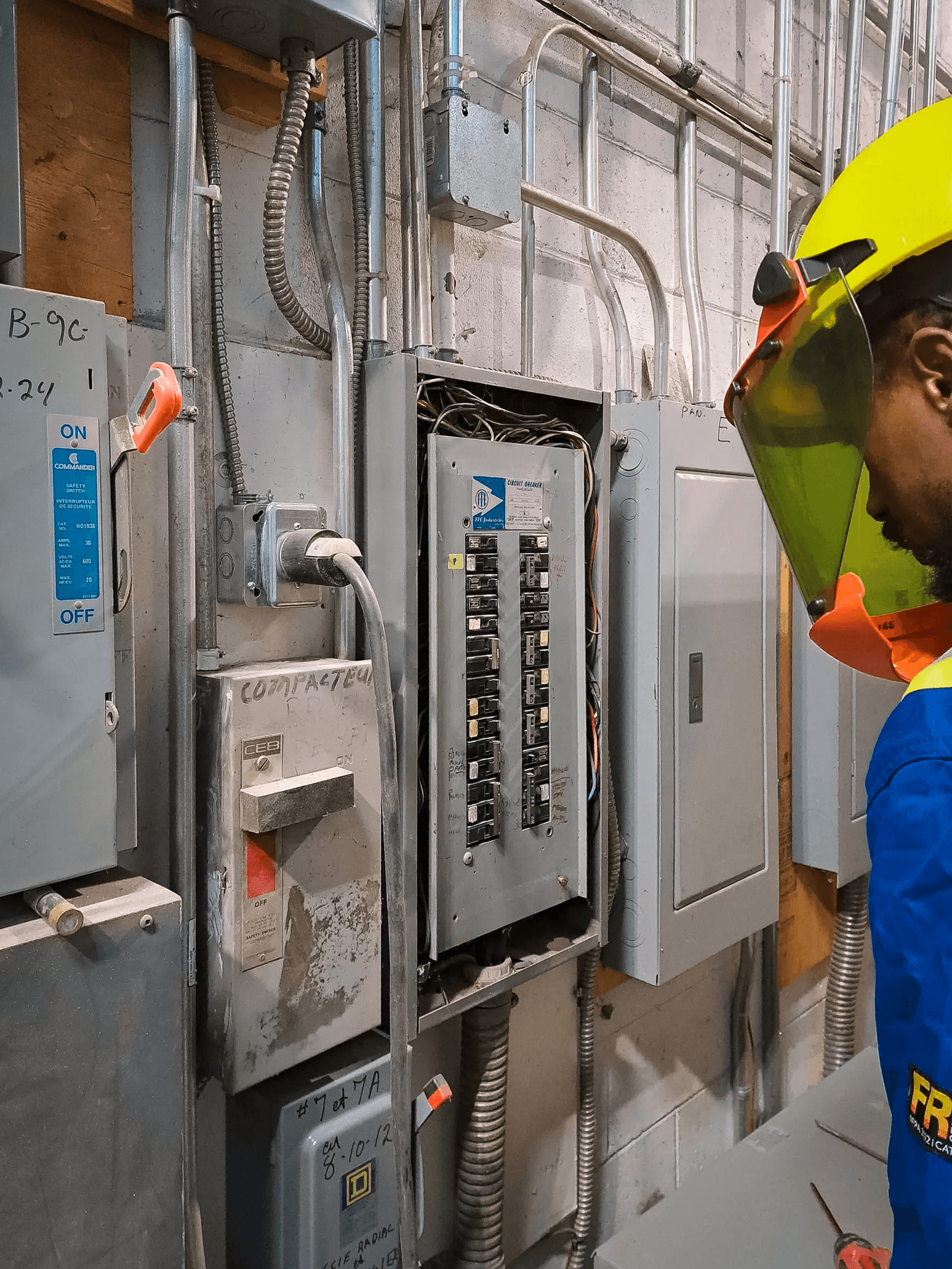 Électricien inspectant un panneau de distribution électrique commercial avec disjoncteurs, conduits métalliques et équipements de protection.