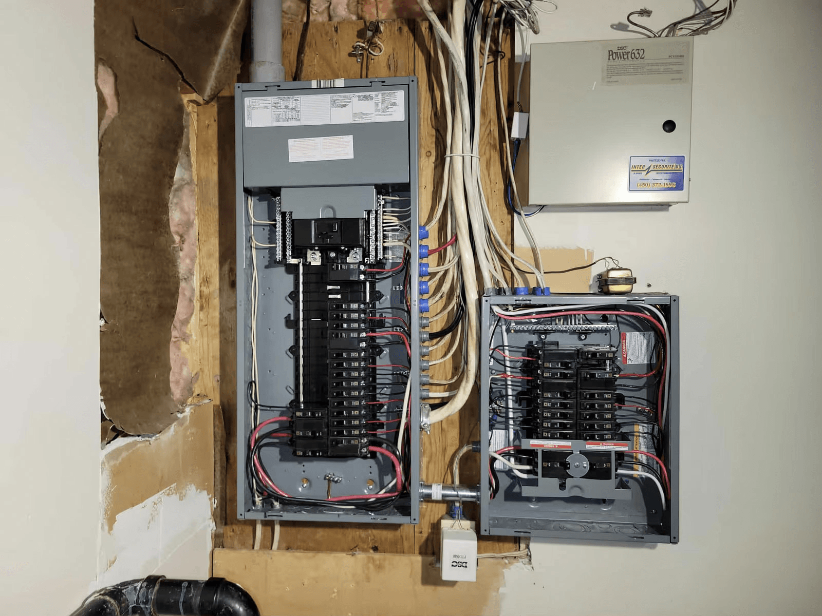 Installation électrique résidentielle avec deux panneaux de disjoncteurs ouverts, câblage apparent et boîtier de système de sécurité fixé au mur.