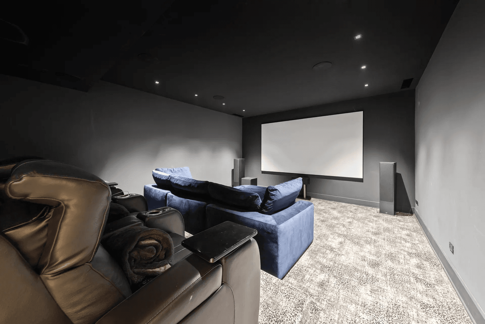 Salle de cinéma maison dédiée, équipée d'un grand écran de projection, d'enceintes, de fauteuils inclinables, d'un canapé et d'un éclairage encastré, le tout dans une pièce sombre.

