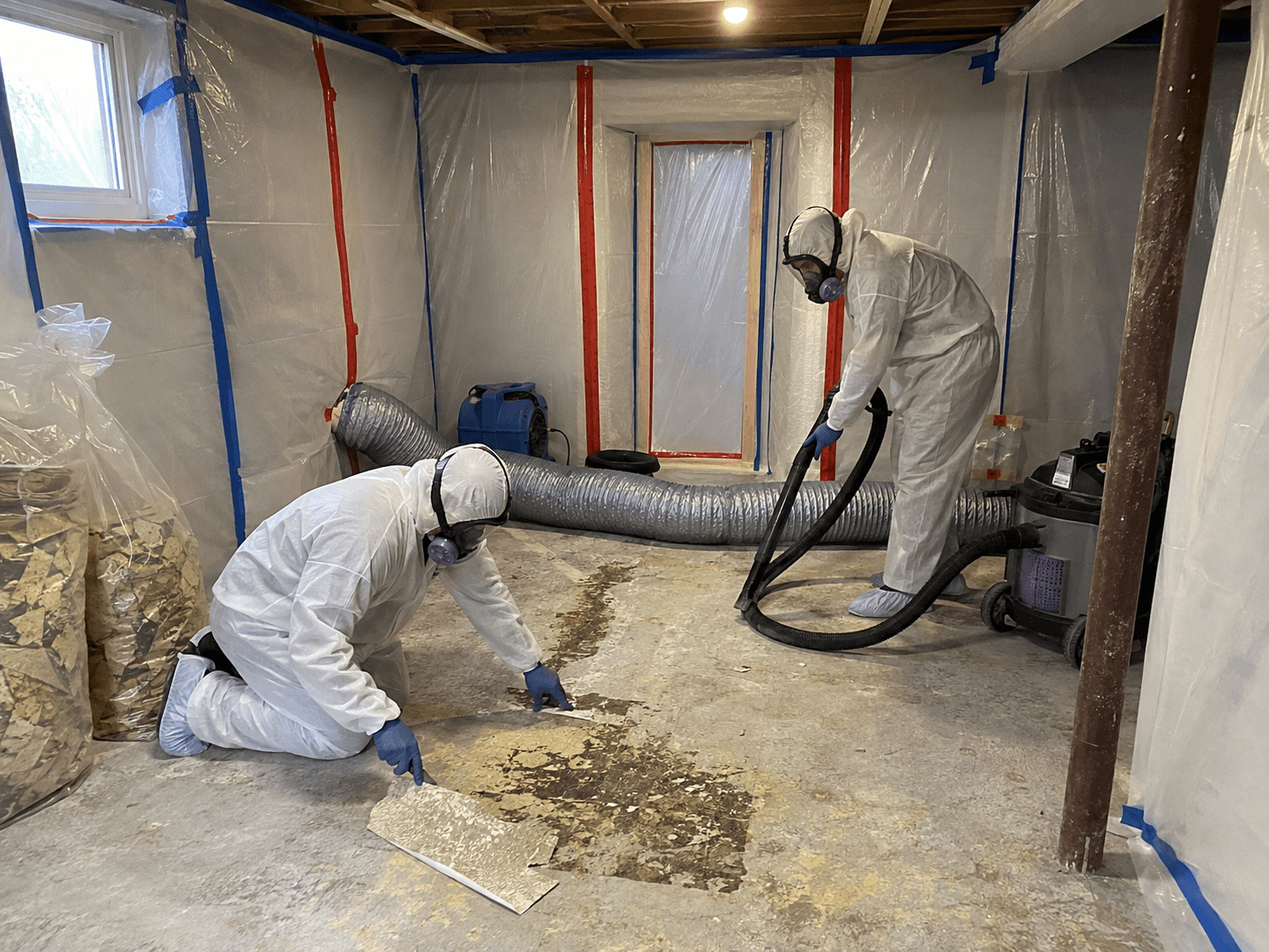 Travailleurs en combinaison de protection retirant un ancien revêtement de sol dans une zone confinée avec bâches de plastique, ventilation et aspirateur industriel / Workers in protective suits removing old flooring inside a contained work area with plastic sheeting, ventilation and an industrial vacuum