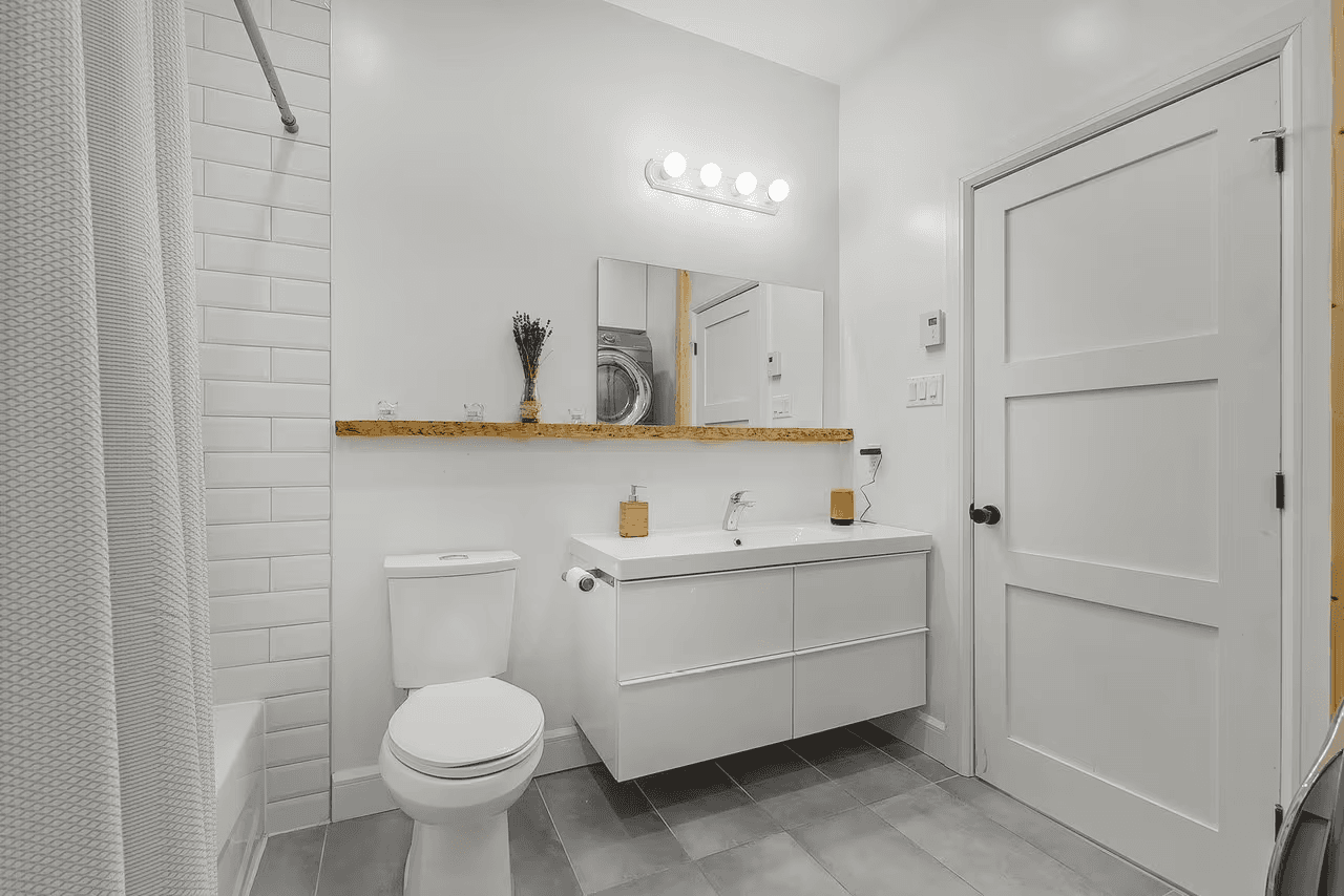 Salle de bain moderne et épurée avec meuble-lavabo blanc suspendu, miroir mural, toilette, douche carrelée, plancher gris et porte intérieure blanche&nbsp;
