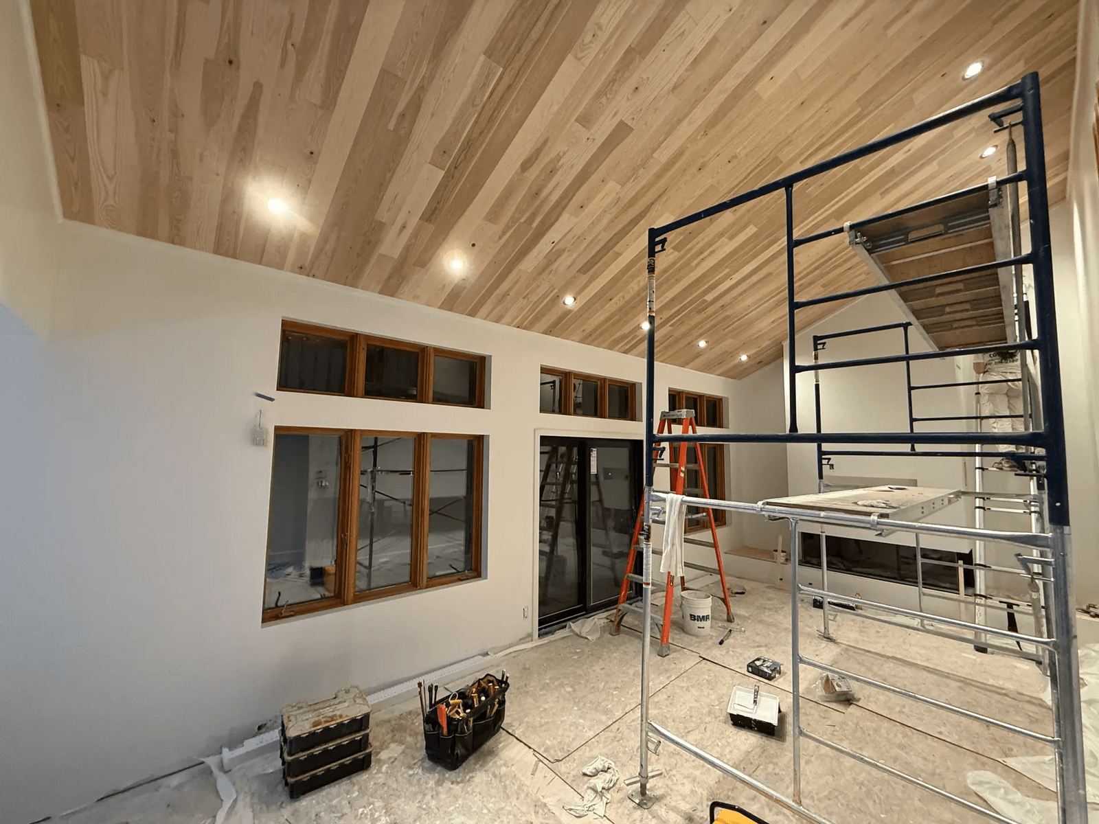 Rénovation intérieure en cours avec plafond cathédrale en lattes de bois, éclairage encastré, grandes fenêtres, portes vitrées, échafaudage et outils de chantier