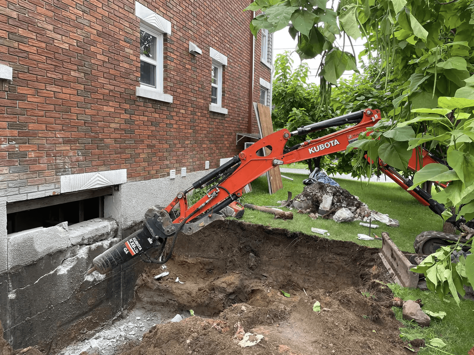 Mini-excavatrice Kubota équipée d’un marteau hydraulique réalisant des travaux d’excavation et de démolition près de la fondation d’une maison en brique.