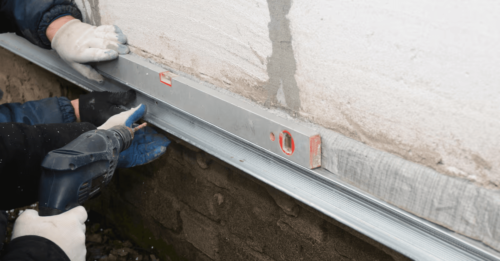 Installation d’un profilé métallique sur un mur extérieur en crépi avec perceuse et niveau à bulle lors de travaux de rénovation.