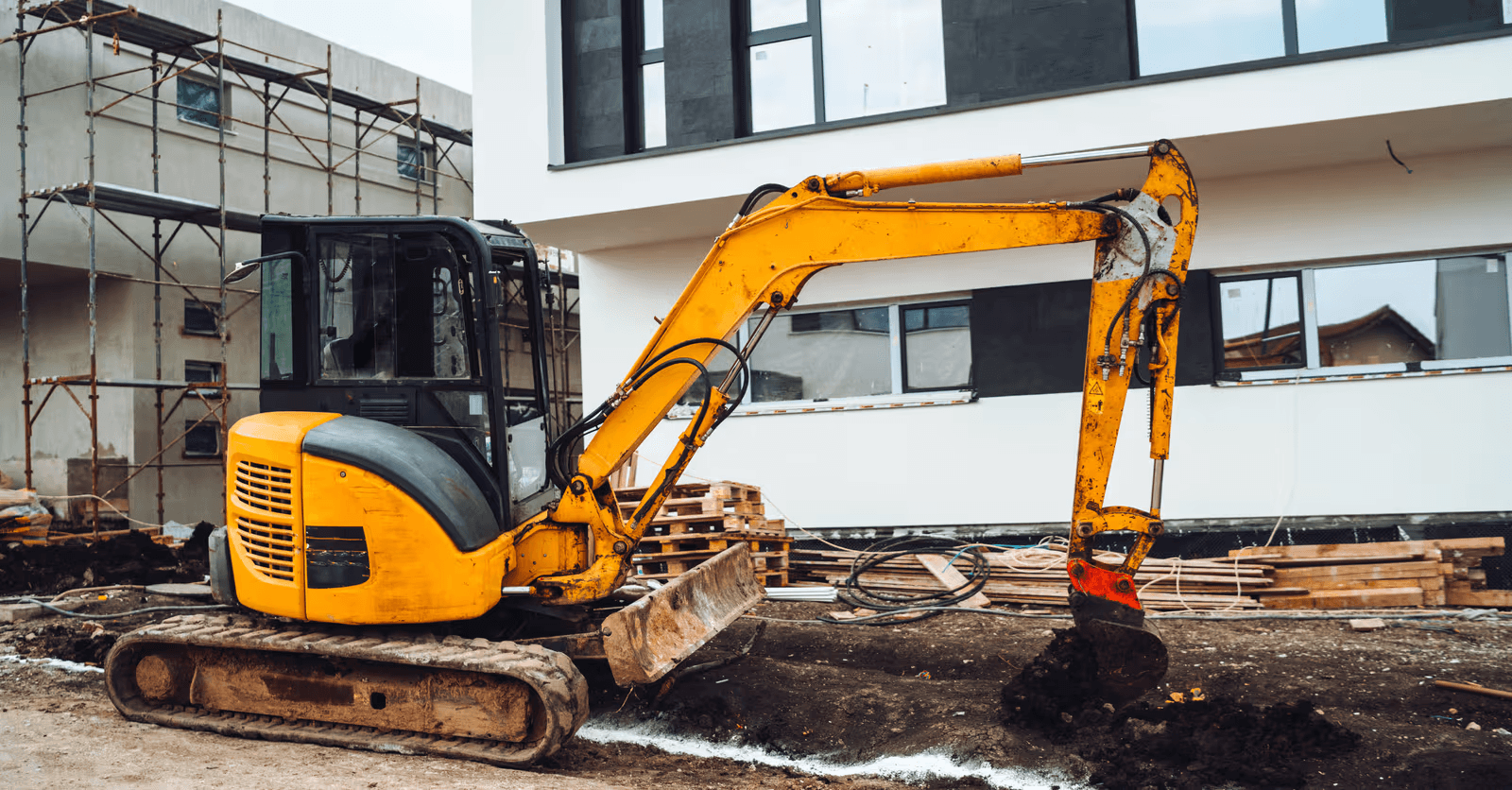 Mini-excavatrice sur chenilles effectuant des travaux de terrassement devant un bâtiment résidentiel moderne en construction, avec échafaudage et matériaux de chantier.