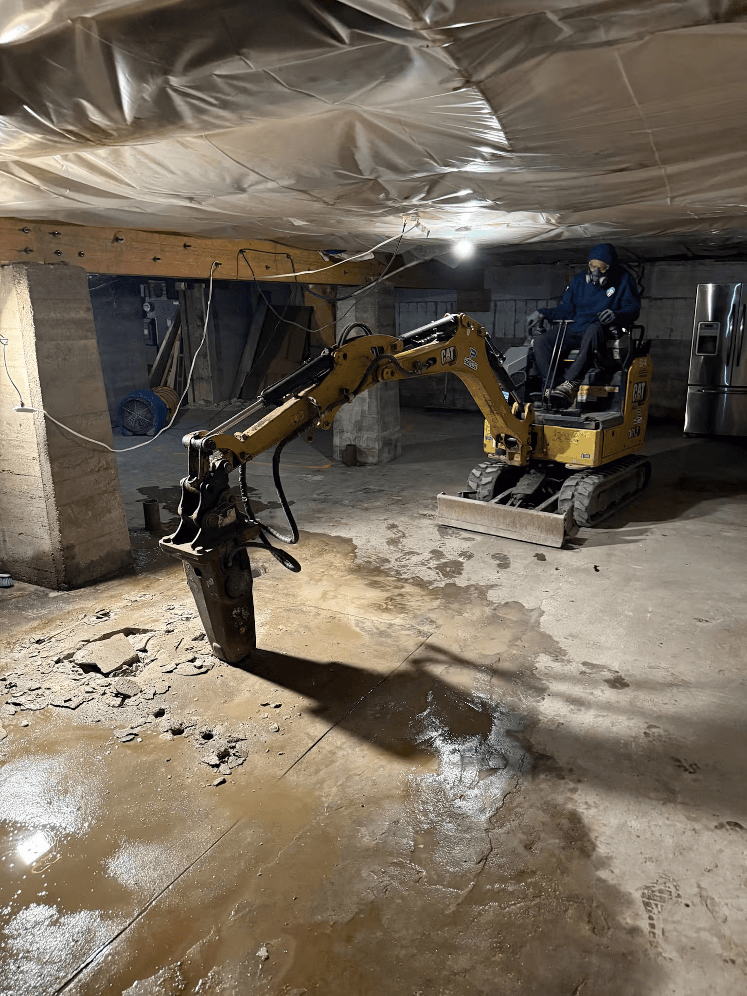 Mini-excavatrice équipée d’un marteau hydraulique démolissant une dalle de béton dans un sous-sol résidentiel en rénovation.