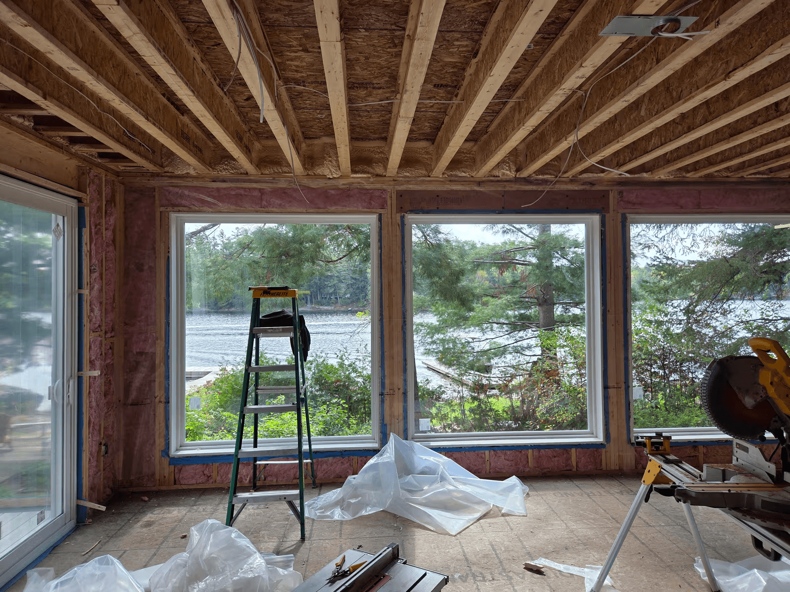 Pièce résidentielle en rénovation avec charpente apparente, isolation en fibre de verre, grandes fenêtres donnant sur l’eau et outils de chantier.