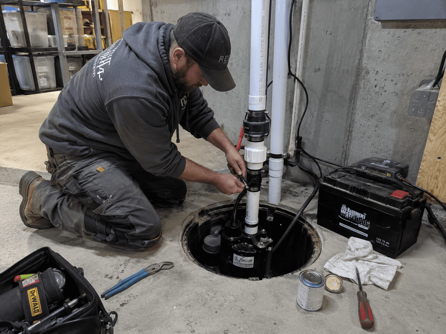 Technicien installant et ajustant une pompe de puisard dans un sous-sol, avec tuyauterie en PVC, batterie de secours et outils de plomberie
