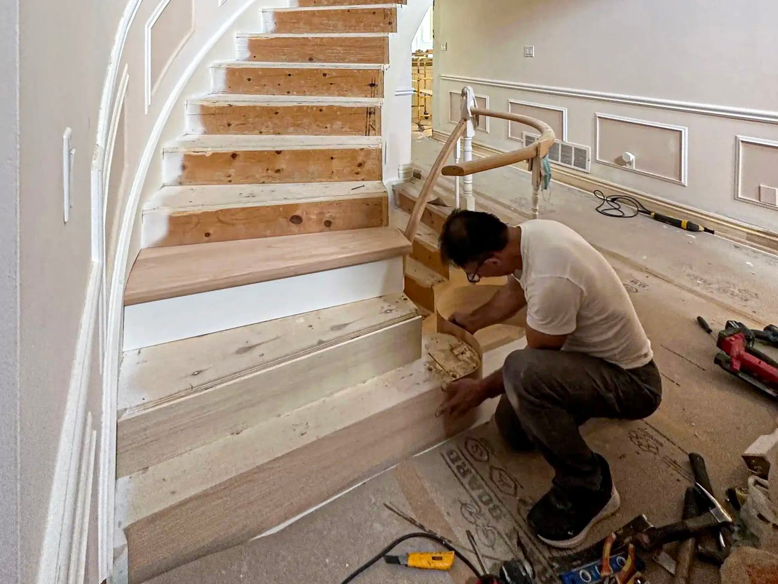 Escalier en bois courbé en cours de rénovation, avec un artisan travaillant sur les finitions dans un intérieur classique avec moulures murales.
