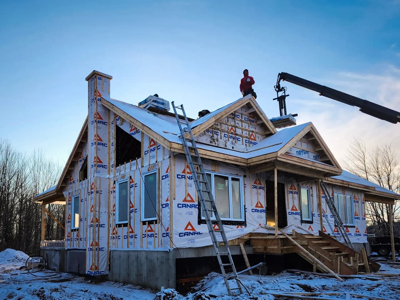Maison en construction en hiver avec revêtement pare-intempéries CANAC, échafaudages, échelles, et ouvriers sur le toit sous un ciel bleu.