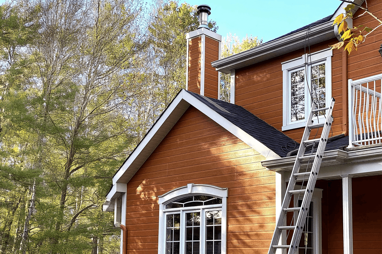 Maison à deux étages avec un revêtement extérieur en bois orange et une cheminée, équipée d'une échelle en aluminium appuyée contre le toit en bardeaux d'asphalte