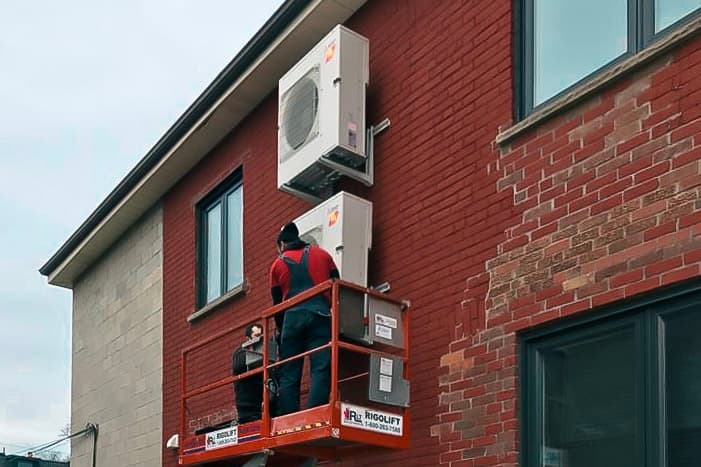 Deux techniciens effectuent l'installation d'unités extérieures de pompes à chaleur sur le mur en briques d'un bâtiment résidentiel, en utilisant une nacelle élévatrice pour un travail en hauteur sécurisé et précis.
