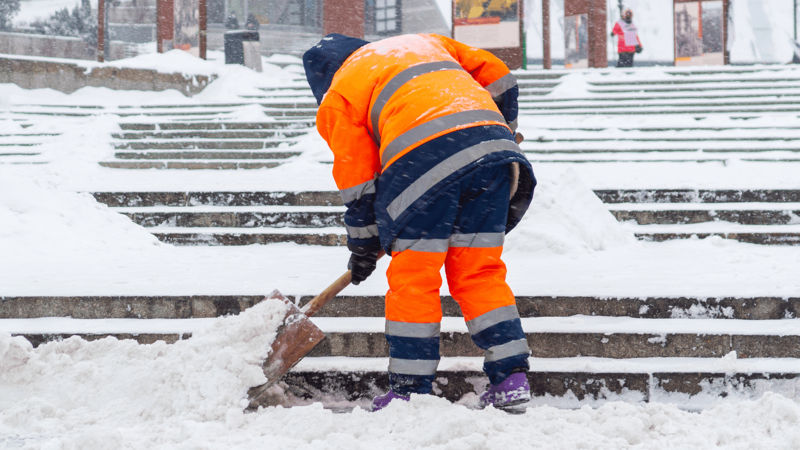 Travailleur en tenue haute visibilité utilisant une pelle pour déneiger un escalier par temps hivernal.