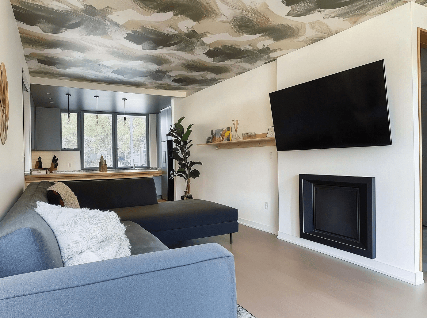 Salon moderne avec canapé sectionnel, télévision murale, foyer encastré noir, plafond artistique et vue sur une cuisine lumineuse avec des fenêtres panoramiques.