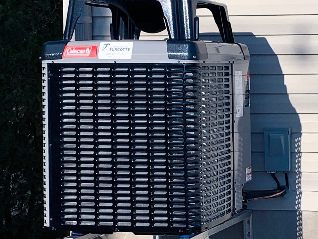 Unité de climatisation extérieure montée sur un support mural en métal à l'extérieur d'une maison avec revêtement en vinyle beige.