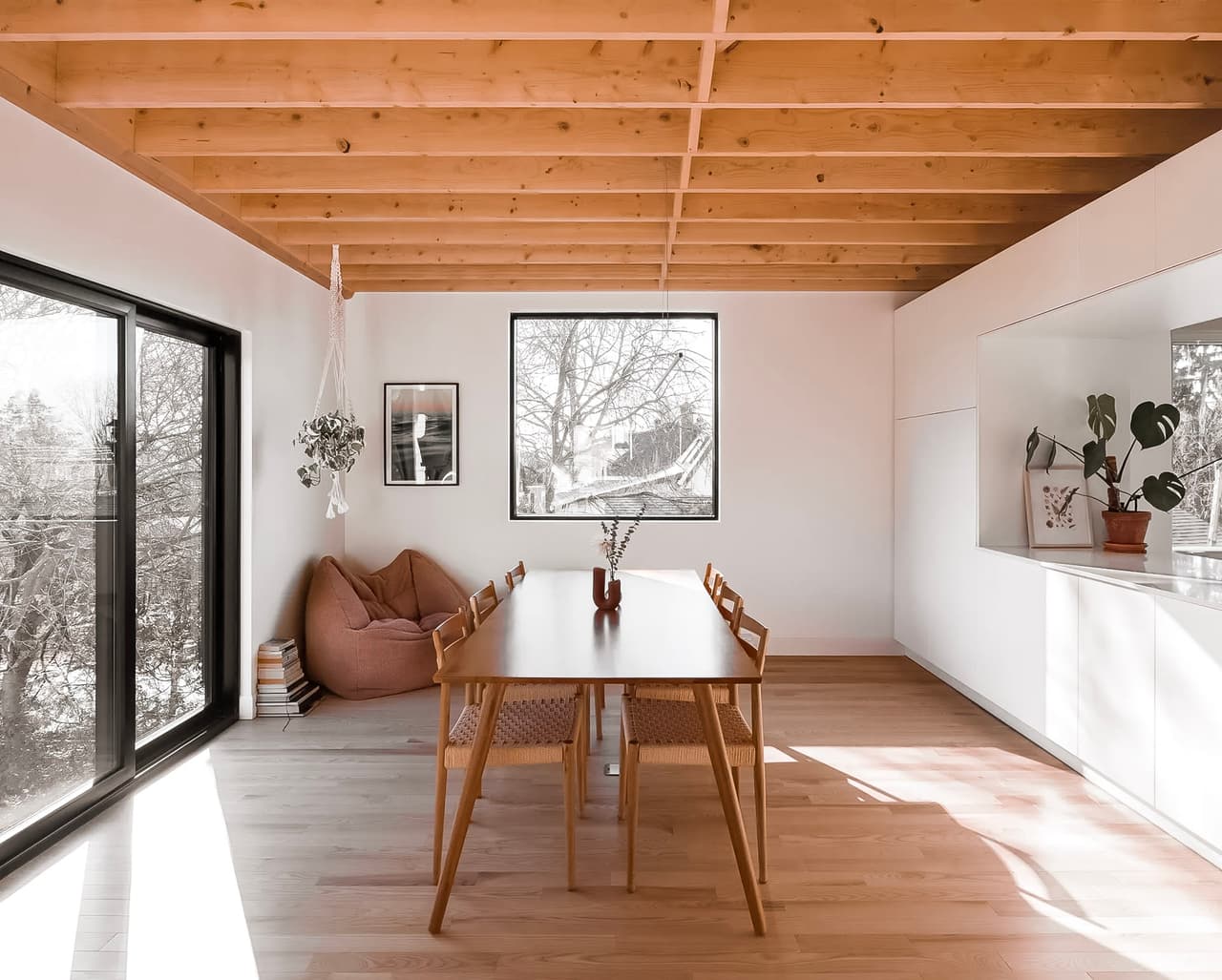 Salle à manger lumineuse avec table et chaises en bois, plafond en poutres apparentes, grandes fenêtres et décoration minimaliste.
