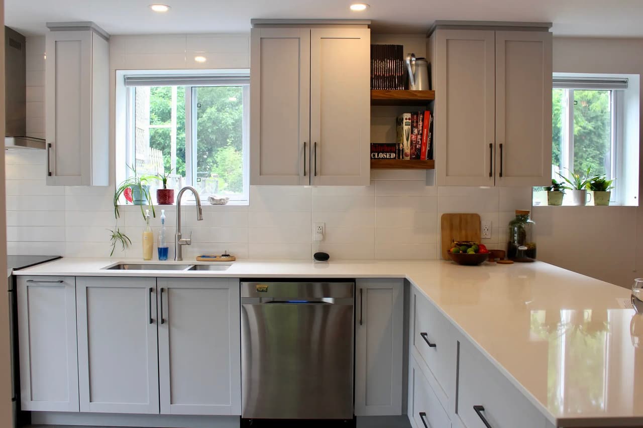 Cuisine contemporaine lumineuse avec armoires grises, comptoir blanc, évier double, lave-vaisselle en acier inoxydable, fenêtres et étagères en bois.