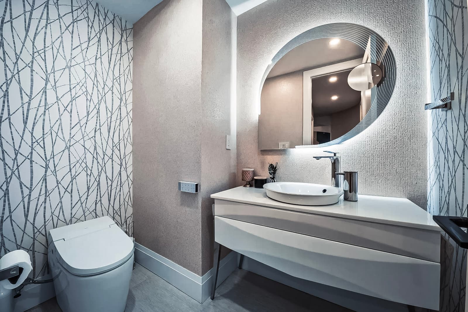 Salle de bain moderne avec toilette intelligente, meuble-lavabo minimaliste blanc, miroir ovale rétroéclairé et mur décoratif à motifs abstraits.