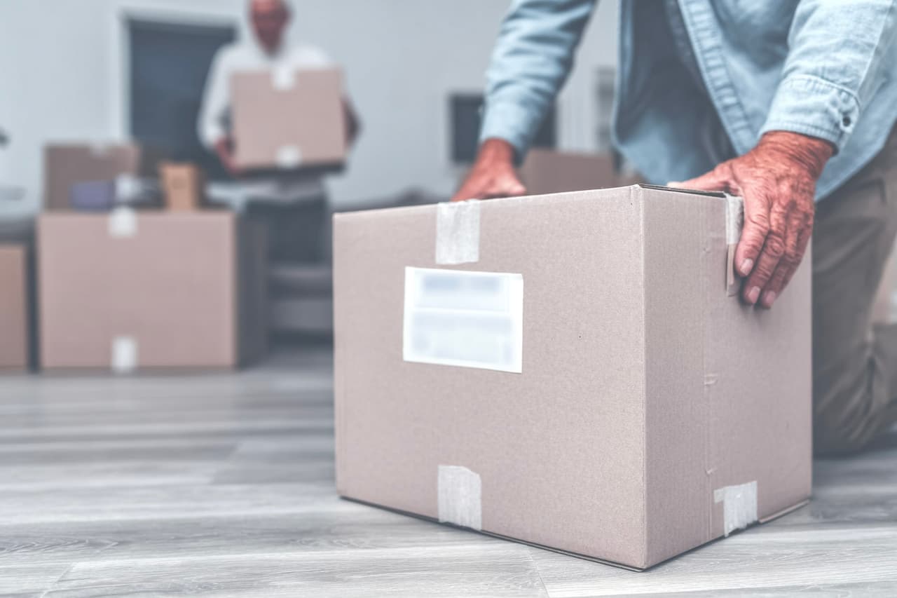 Déménagement résidentiel avec cartons scellés posés sur plancher moderne et homme transportant une boîte dans une maison.