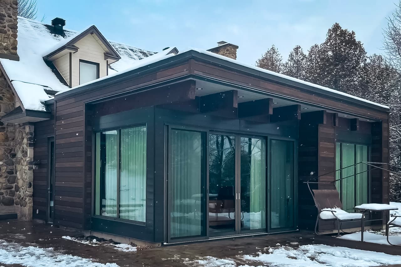 Extension de maison moderne à un étage avec de grandes baies vitrées coulissantes et un revêtement en bois foncé, sous la neige en hiver