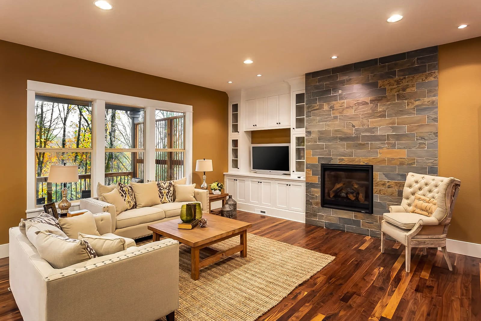 Salon chaleureux avec foyer encastré en pierre, meubles beiges et grandes fenêtres donnant sur une terrasse.