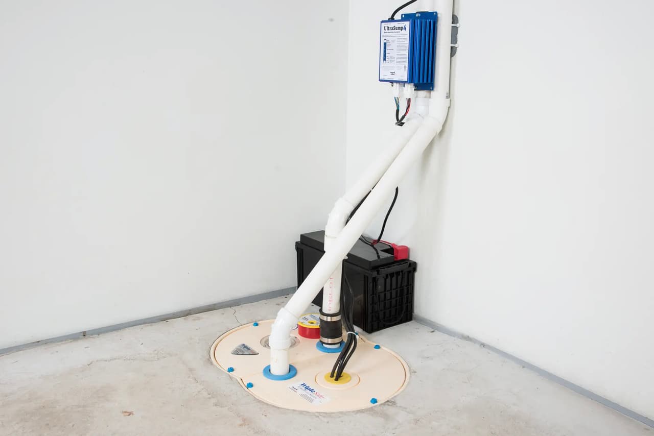 Système de pompe de puisard avec tuyaux en PVC, batterie de secours et boîtier électrique installé dans un sous-sol.