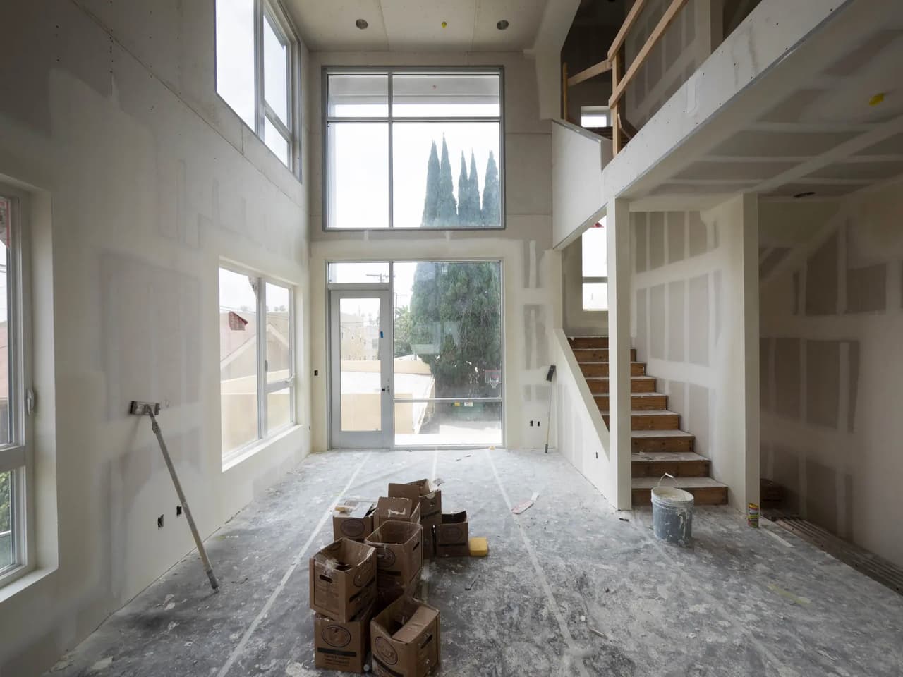 Intérieur de maison en construction avec plafond cathédrale, mezzanine, escalier, murs en gypse et grandes fenêtres laissant entrer une abondante lumière naturelle.