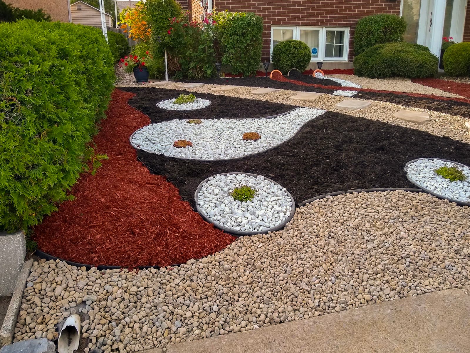 Aménagement paysager inspiré du yin-yang avec paillis coloré, pierres décoratives et plantes succulentes devant une maison en brique.