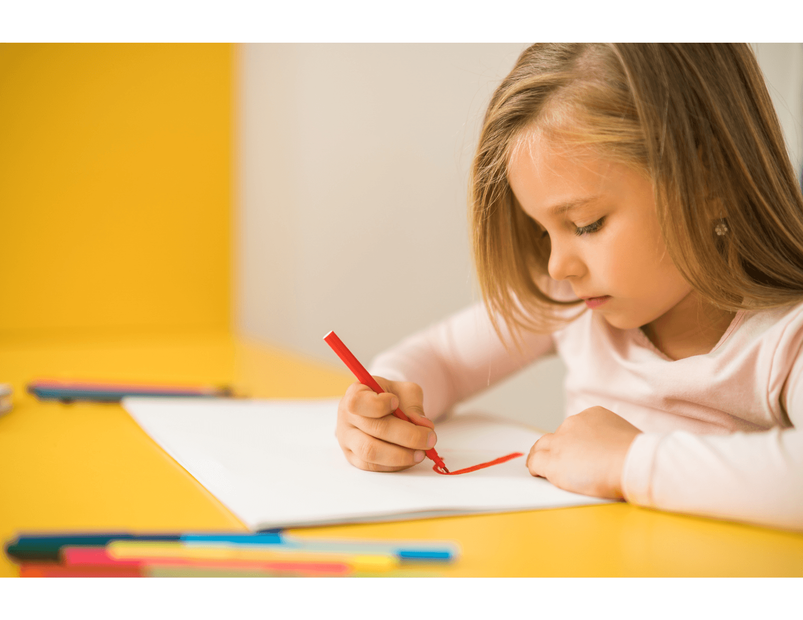 Petite fille dessinant avec un crayon rouge sur une feuille de papier, assise à une table jaune.