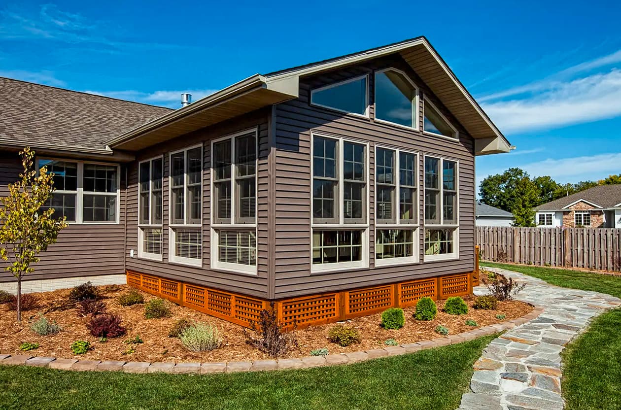 Extension de maison comprenant une grande véranda vitrée, un revêtement extérieur en bois, un aménagement paysager avec paillis et un sentier en pierre, sous un ciel bleu.