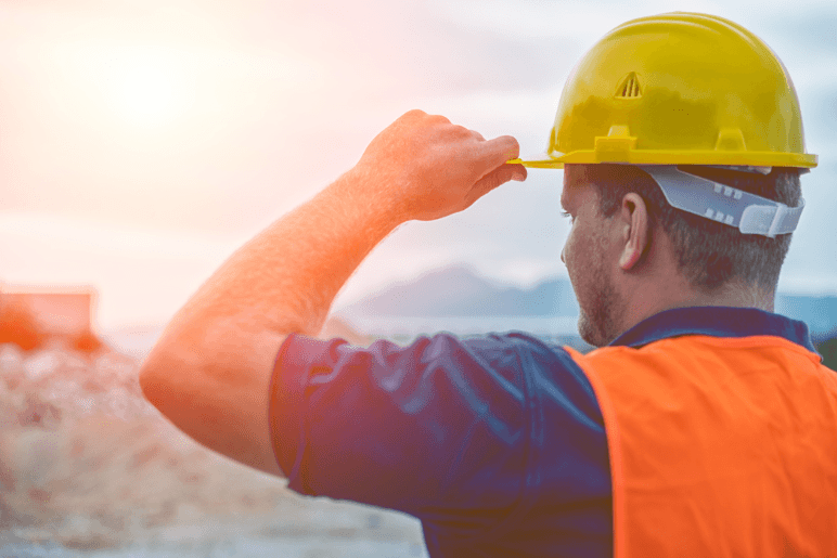 Travailleur de la construction portant un casque jaune et un gilet de sécurité orange, regardant un chantier au lever du soleil, symbolisant la continuité et la vigilance malgré la grève dans le secteur résidentiel.