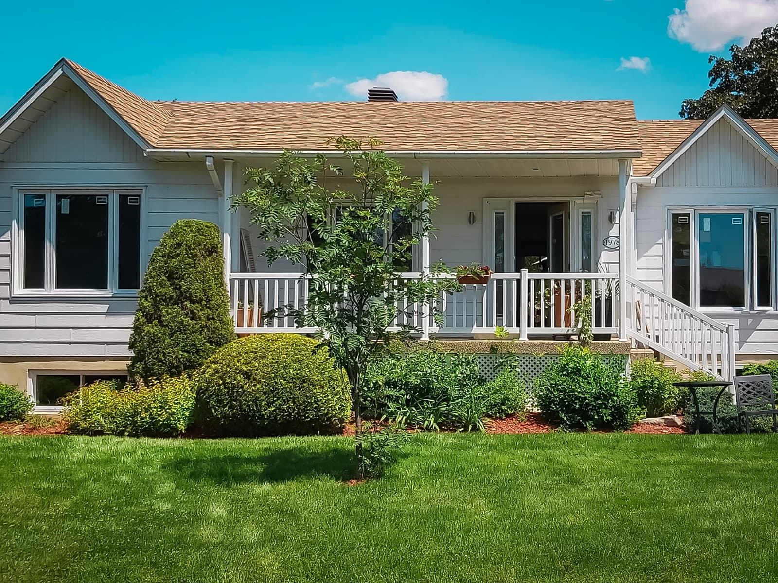  Maison de plain-pied avec terrasse en bois blanc, jardin paysager et pelouse verte sous un ciel bleu clair.