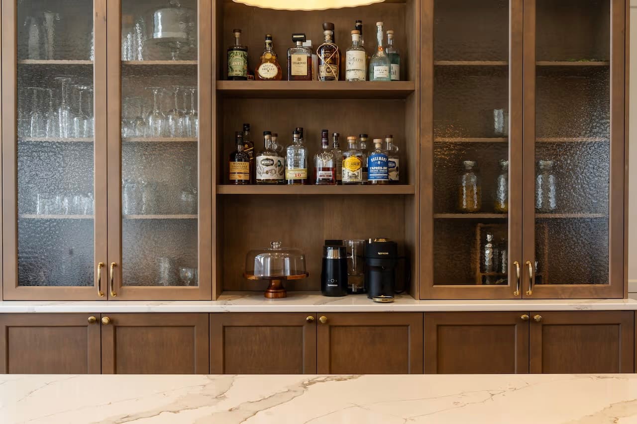 Armoire-bar en bois foncé avec portes vitrées, bouteilles d’alcool et petits électroménagers.