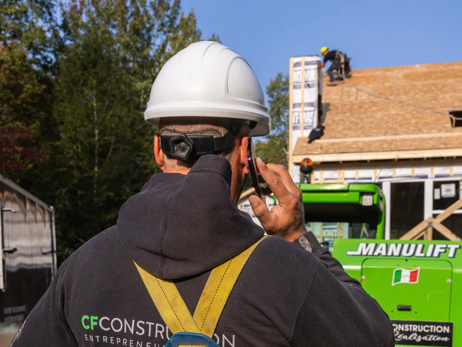 Travailleur de la construction portant un harnais de sécurité et un casque, en communication téléphonique sur un chantier avec des travaux de toiture en arrière-plan.