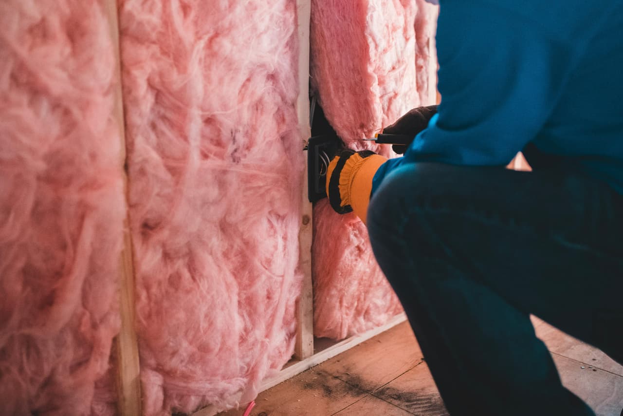 Travailleur effectuant une installation électrique dans un mur résidentiel isolé avec de la fibre de verre rose pendant des travaux de construction ou de rénovation.