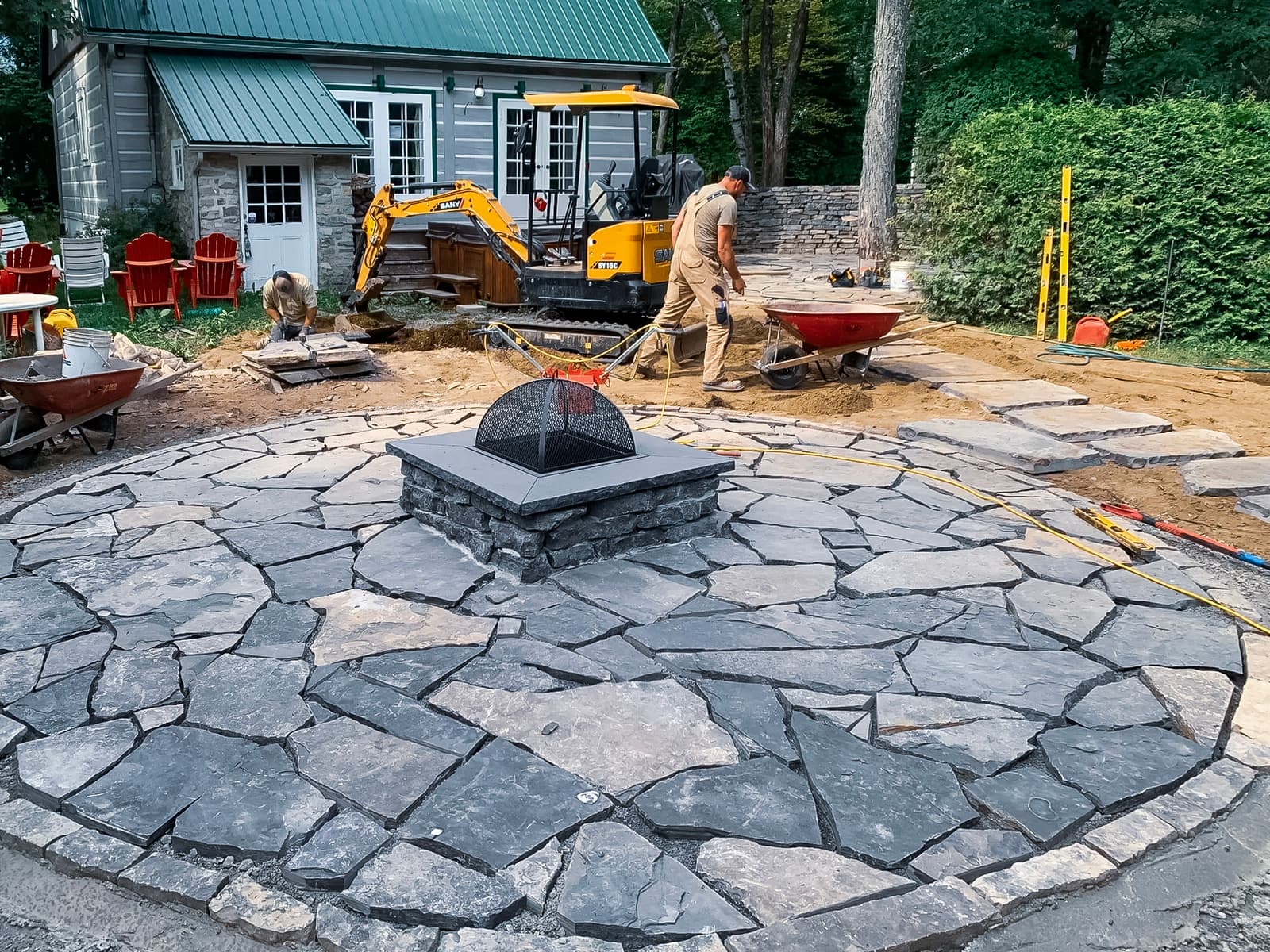 Installation d'un foyer extérieur sur une surface en pierres naturelles dans un aménagement paysager en construction devant une maison en bois et métal vert.