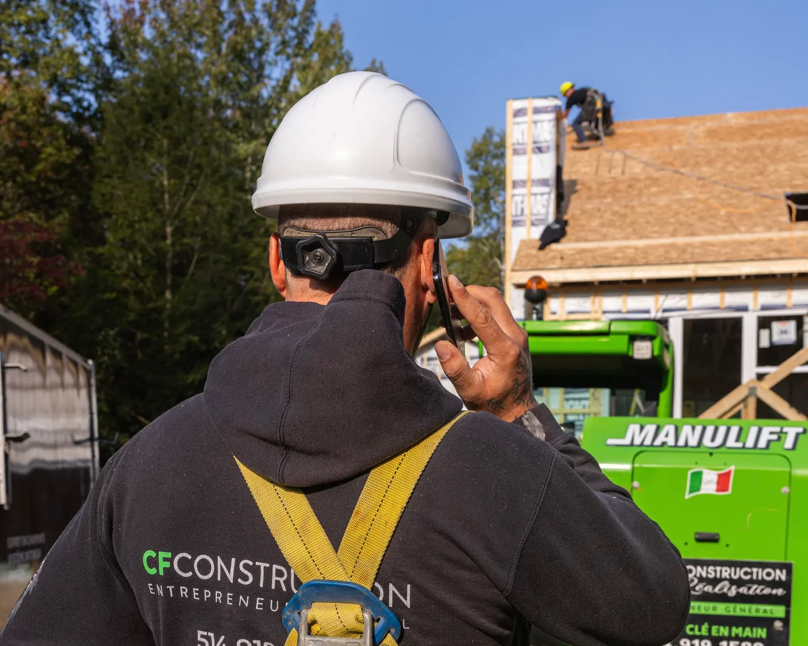 Travailleur de la construction portant un harnais de sécurité et un casque, en communication téléphonique sur un chantier avec des travaux de toiture en arrière-plan.