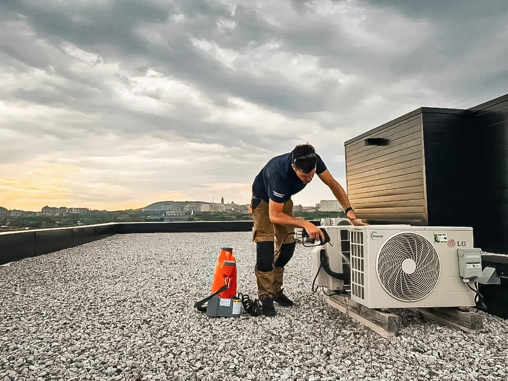 Technicien effectuant l'installation d'une unité extérieure de climatisation sur un toit plat avec vue urbaine.