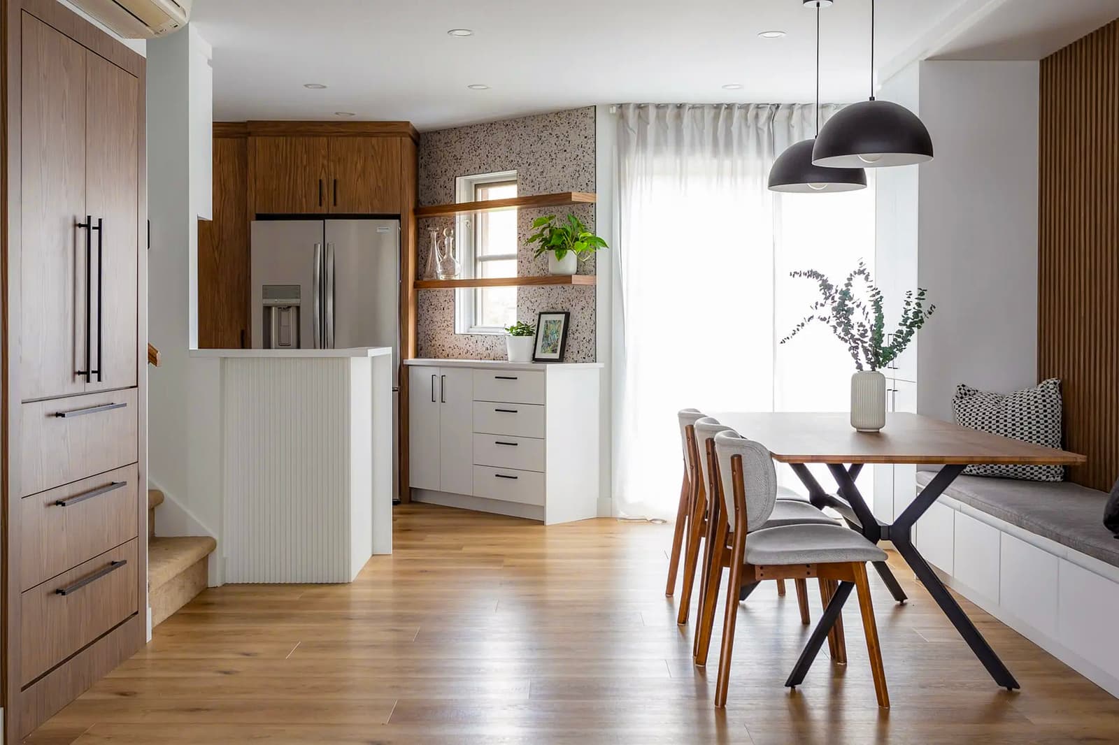 Cuisine et salle à manger modernes avec meubles en bois, table en bois avec chaises rembourrées et banquette intégrée. Plancher en bois clair, éclairage suspendu noir et étagères ouvertes décorées de plantes.